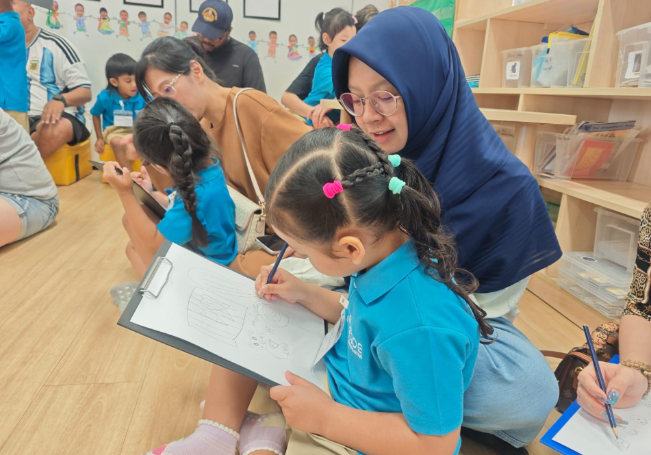 Teacher in hijab helps a girl in blue uniform draw on a clipboard in a classroom with other children.